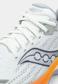 Bílé běžecké boty s prodyšnou síťovinou, šedými akcenty a jasně oranžovou podrážkou. Obsahují výrazné logo a design s kulatým vzorem.