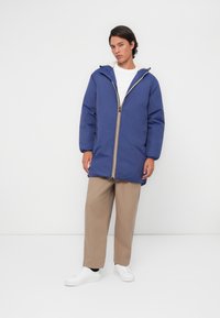 Blå isolert jakke med glidelås foran, hette og gule detaljer. Kombinert med beige vide bukser og hvite joggesko, satt mot en enkel bakgrunn.