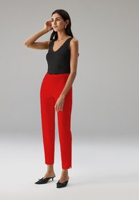 Zwarte geribbelde tanktop met een V-hals, gecombineerd met strakke rode broeken met scheuren bij de enkels, en zwarte puntige muiltjes.