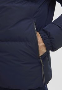 Marineblaue Steppjacke mit einer Reißverschlusstasche, gerippten Bündchen und einer glatten, isolierten Textur. Verfügt über ein metallisches Reißverschluss-Detail an der Tasche.