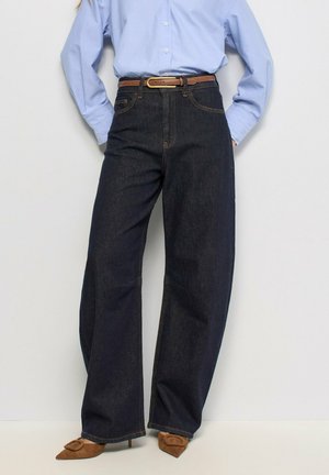 Persoon die donkerblauwe, hoog taille, wijde pijpen jeans draagt met een bruine riem, lichtblauw overhemd met knopen en bruine puntige hakken.