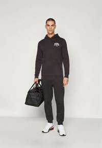 Felpa nera con cappuccio e tasca a canguro, logo sul petto, abbinata a pantaloni della tuta neri, scarpe da ginnastica bianche e una borsa a tracolla nera.