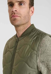 Olijfgroene gewatteerde vest met een staande kraag en rits, voorzien van een gestructureerde binnenlaag en een subtiel golvend stiksels patroon.