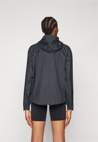 Veste noire légère avec capuche, en tissu texturé et avec des poignets élastiques. Portée par-dessus un short noir, vue de dos.