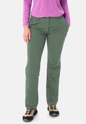 Donna che indossa pantaloni da trekking verde oliva con cerniera e scarpe da trekking marrone chiaro, in piedi con le mani nelle tasche su sfondo bianco.