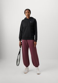 Svart hettegenser med snor, kombinert med løse bordeaux joggebukser og hvite joggesko. Holder et tennisslag i den ene hånden.
