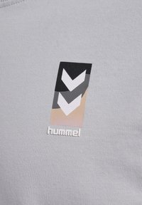 Logo Hummel con doppie chevron bianche su rettangoli neri e grigi sopra uno sfondo beige su tessuto grigio chiaro.
