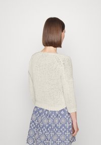 Strickpullover in Off-White mit lockerer Passform, Dreiviertelärmeln und einem strukturierten Muster. Kombiniert mit einem blauen Rock mit weißer Stickerei.