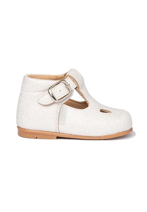 Zapato infantil blanco brillante con tira en el tobillo y hebilla plateada, suela marrón claro y detalle recortado en la parte delantera.