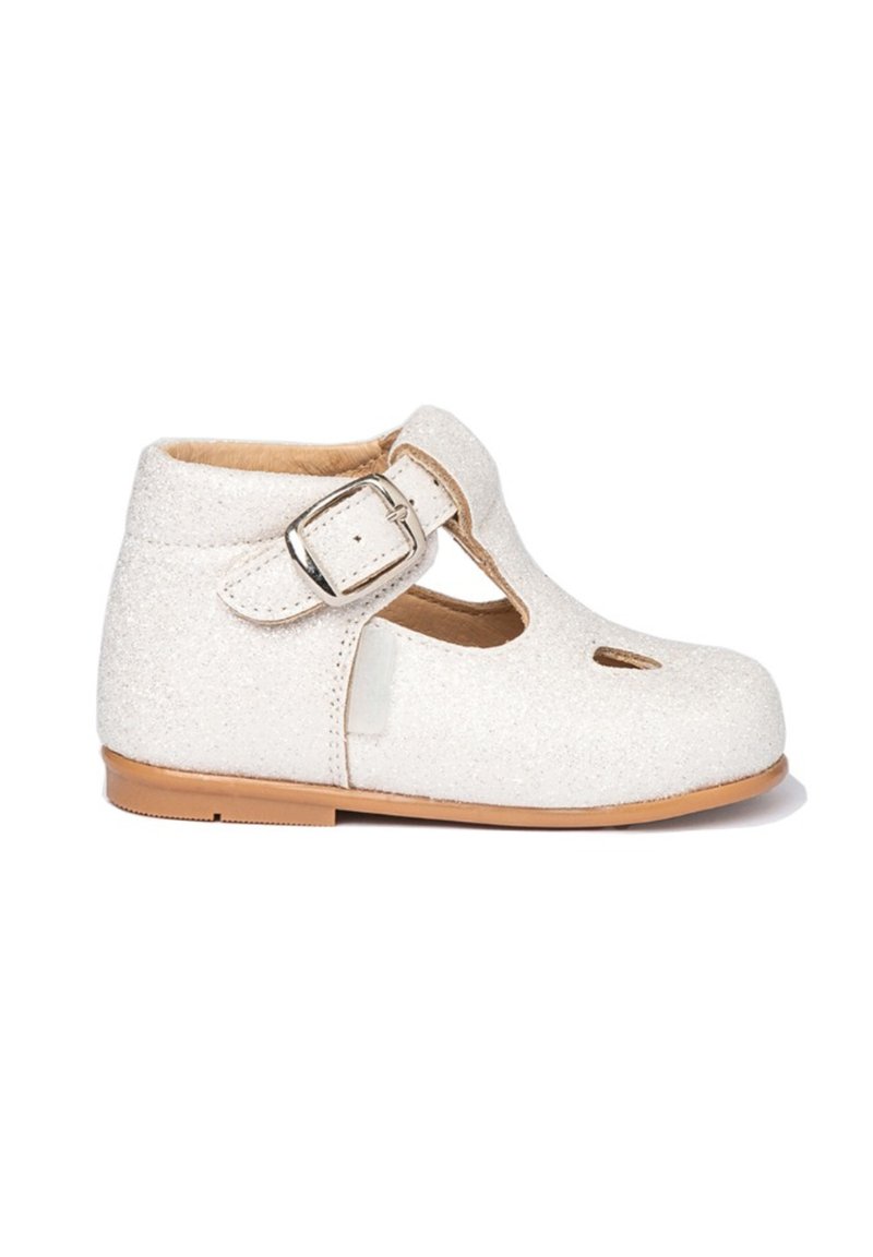 Zapato infantil blanco brillante con tira en el tobillo y hebilla plateada, suela marrón claro y detalle recortado en la parte delantera.