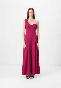 Fuchsia, en-skulder jumpsuit med plisserte vide ben og strukturert liv. Glatt materiale med matt finish, kombinert med svarte sandaler.