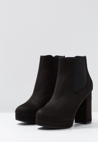 Botines de ante negro con tacón cuadrado, que presentan paneles laterales elásticos, punta redondeada y un diseño sin costuras. Textura suave en toda la superficie.