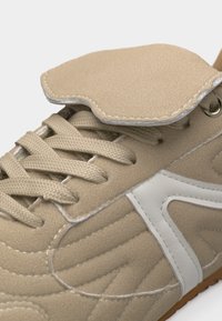 Μπεζ αθλητικό παπούτσι με κορδόνια στο ίδιο χρώμα, λευκές λεπτομέρειες στο πλάι και επενδεδυμένη γλώσσα που καλύπτει μερικώς τα κορδόνια.