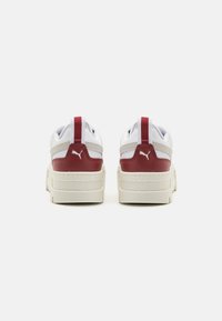 Weiße Sneakers mit roten Akzenten an der Ferse, strukturierten Gummisohlen und weichen Obermaterialien aus Stoff. Mit einem auffälligen Markenlogo an der Seite und auf der Rückseite.