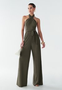 Khaki jumpsuit with a halter neckline, tied waist, wide legs, textured fabric, and a subtle sheen, paired with a white handbag.