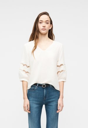 Naine, kes kannab valget V-kaelusega pluusi pitsiliste lõigetega puffvarrukatel ja siniseid teksapükse musta vööd kinnitades, seisab ühevärvilise tausta ees.