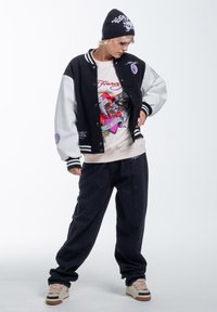 Black and grey varsity jacket with embroidered patches, white t-shirt featuring colorful graphic design, dark denim pants, and beige sneakers.