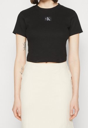 Osoba nosząca czarny krótki t-shirt Calvin Klein i białą spódnicę z wysokim stanem, pokazująca tatuaże na obu przedramionach.
