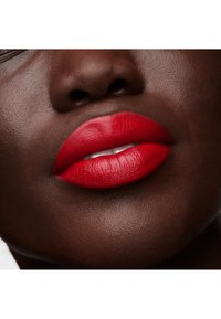 Czerwona, intensywna szminka na pełnych ustach, podkreślająca gładką, matową teksturę na tle głębokiej brązowej skóry. Subtelny połysk wzdłuż linii ust.
