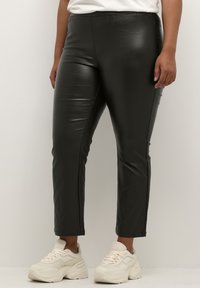 Schwarze Leggings aus Kunstleder mit schlanker Passform, die eine glatte Textur aufweisen und keine sichtbaren Verschlüsse haben, kombiniert mit hellen Sneakers.