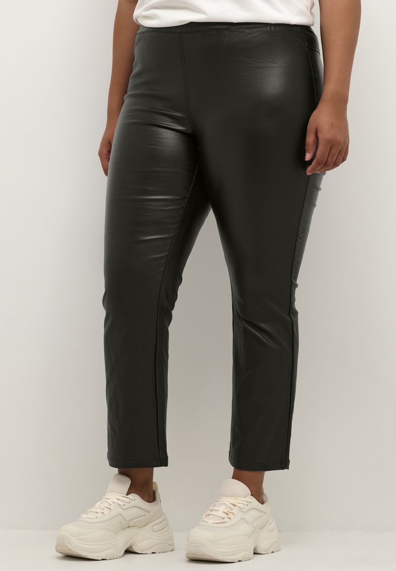 Schwarze Leggings aus Kunstleder mit schlanker Passform, die eine glatte Textur aufweisen und keine sichtbaren Verschlüsse haben, kombiniert mit hellen Sneakers.