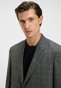 Blazer grigio a quadri con un motivo testurizzato e rever affilati, indossato sopra una camicia nera. Il tessuto appare su misura e strutturato.