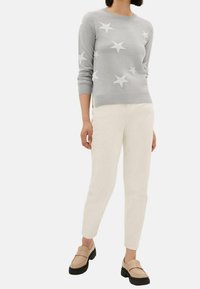 Femme portant un pull gris clair avec des étoiles blanches, un pantalon blanc cassé et des mocassins beiges à semelles noires, debout devant un fond uni.