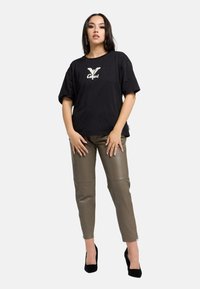 Schwarzes Baumwoll-T-Shirt mit weißem Logo, kombiniert mit olivgrünen Lederhosen. Die Schuhe sind schwarze High Heels. Schlichtes, modernes Design.