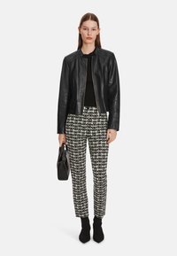 Schwarze Lederjacke mit Reißverschluss, schwarzer Pullover, gemusterte weiße und schwarze Hose und schwarze Knöchelstiefel, hält eine kleine schwarze Tasche.