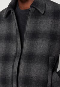 Man som bär en mörkgrå och svart rutad ull zip-up kappa över en svart ribbad crewneck-tröja, visad från nacke till midja.