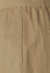 Beige katoenen stof met een zachte textuur, voorzien van een zijzak en subtiele plooien bij de tailleband. Eenvoudig en functioneel ontwerp.
