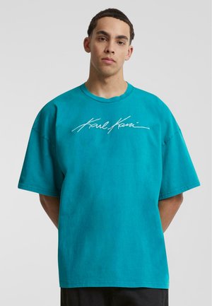 Tirkīza krāsas oversized T-krekls, kas izgatavots no kokvilnas, ar baltu izšūtu logo "Karl Kani" priekšpusē un apaļu kakla dizainu.