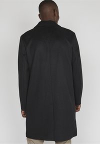 Uomo che indossa un lungo cappotto nero, rivolto di spalle, in piedi contro uno sfondo grigio chiaro uniforme.