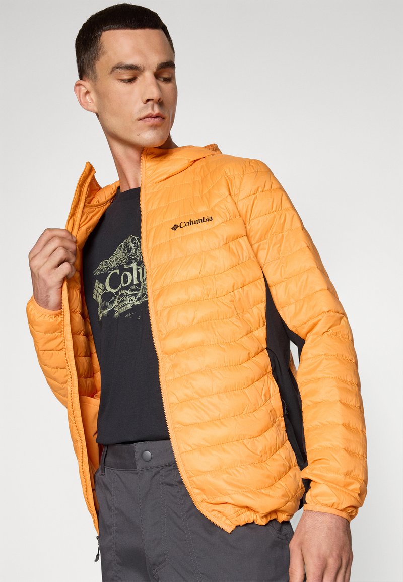 Veste isolée orange avec un design matelassé, fermeture éclair à l'avant, accents noirs et un logo sur la poitrine, portée sur un t-shirt graphique noir.