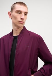 Burgundijas bomber jakas ar ribotu apkakli un aprocēm, ar rāvējslēdzēja aizdari un maigu logo detaļu uz krūtīm; gluda, viegla auduma.