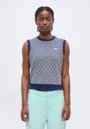 Noor naine punutud juustega kannab varrukateta tumesinist mustrilist topi ja helemintrohelisi kõrge vöökohaga pükse, seisab ühevärvilise taustaga.