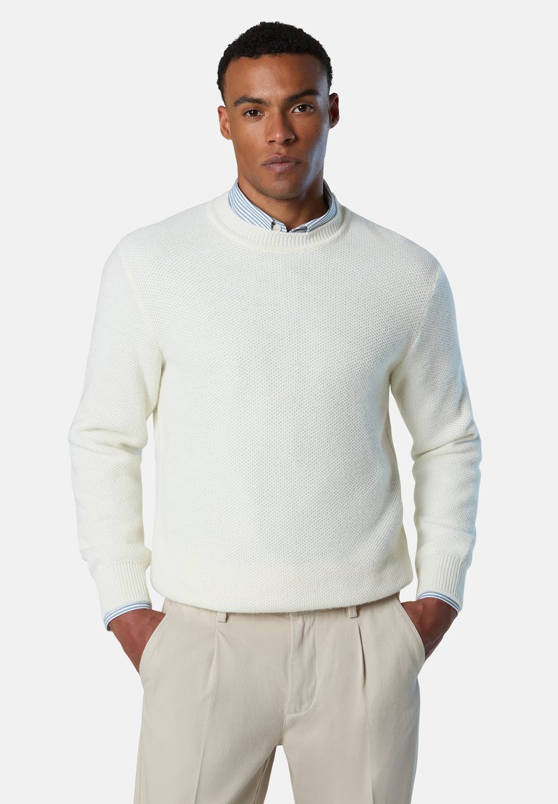 Cremig strukturierter Strickpullover mit einem gerippten Rundhalsausschnitt und Bündchen, getragen über einem hellblauen und weißen gestreiften Hemd. Die Hose ist beige.