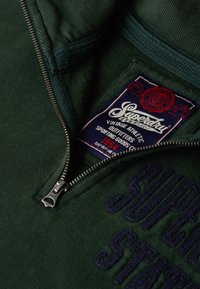 Zielona bluza z ćwierć zamkiem, miękka w dotyku, z ciemnoniebieskim haftowanym logo i etykietą w stylu vintage wewnątrz.
