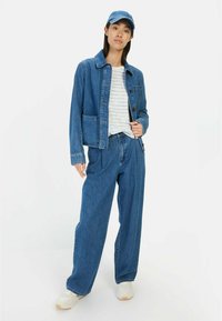 Denimjacket met twee voorzakken, gecombineerd met loszittende denim broek en een gestreept wit-blauw shirt. Een blauwe pet en sneakers maken de look compleet.