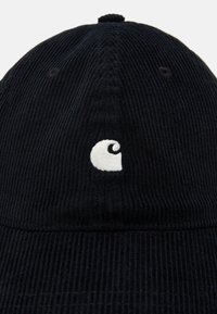 Svart kordfløyelshette med hvit brodert logo, avrundet krone og en teksturert overflate med synlige sømdetaljer.