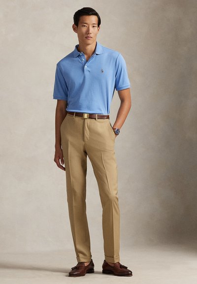 Polo azul claro com gola, calças de fato bege, sapatos de pele castanhos e um relógio. Tecido macio com corte ajustado.