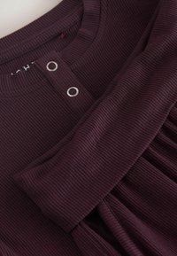 Top ribbed de culoare vișinie, cu decolteu rotund, două accente cu butoane argintii și o textură plisată. Material neted cu un ușor elastic.