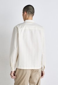 Camicia button-up beige chiaro con righe verticali, colletto classico e maniche lunghe. Tessuto morbido con una vestibilità rilassata, abbinata a pantaloni beige.