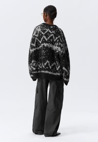 Pull surdimensionné noir et gris à motifs avec une texture en tricot, présentant des designs géométriques et un ourlet côtelé. Associé à un pantalon cargo foncé.