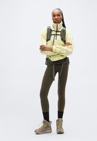 Ελαφρύ κίτρινο fleece μπουφάν με σχέδια, κολάν ελιάς και μπεζ ορειβατικά μποτάκια. Διαθέτει θώρακα με ιμάντες και ζώνη για αποθήκευση.