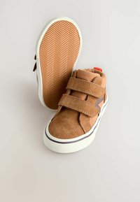 Brune ruskind højtop sko med to Velcro remme, hvid gummisål og herringbone tekstureret ydersål. Har en rød accentstrop.