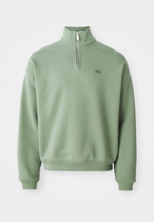 Grønn fleecepuls med kvart glidelås foran, lange ermer, ribbestrikkede mansjetter og en liten brodert logo på brystet.