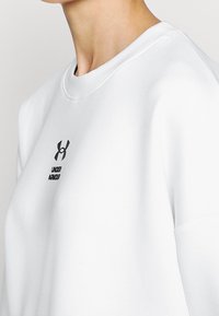 Bílá mikina s kulatým výstřihem, na hrudi s černým logem a textem. Jednoduchá, hladká látka s volnějším střihem.