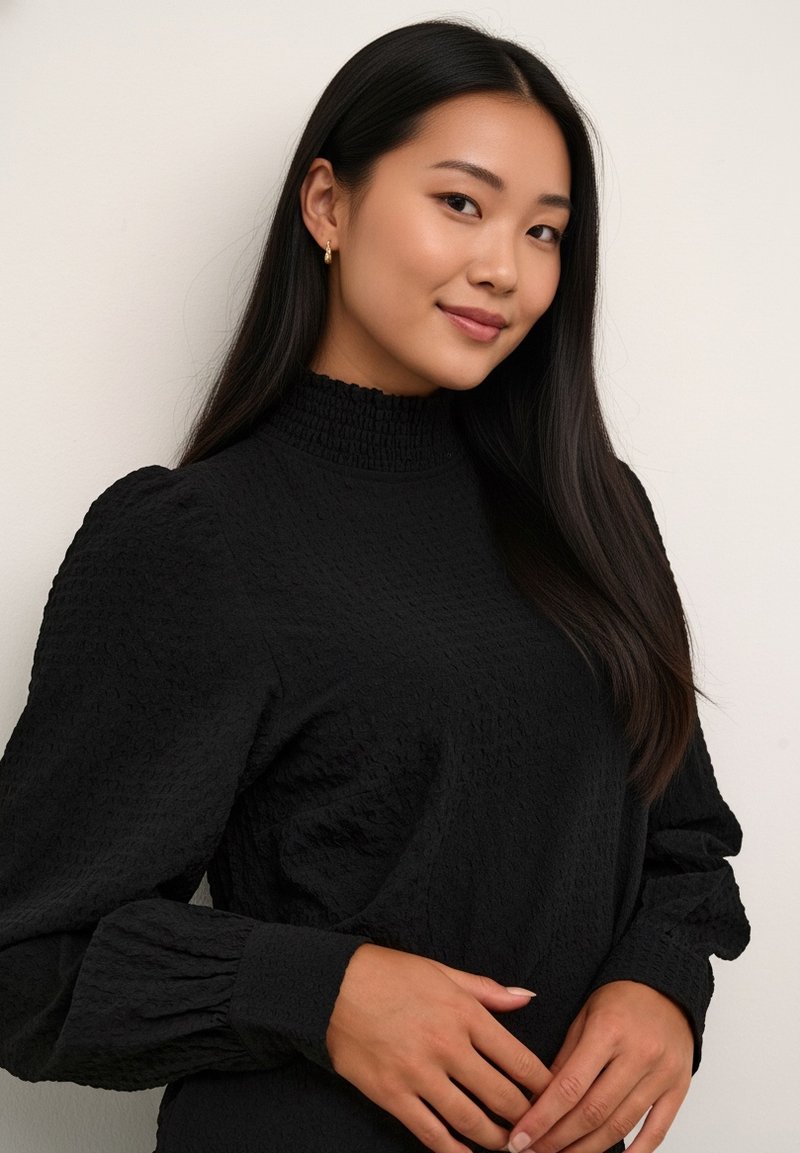 Haut noir à manches longues avec un tissu texturé, col montant et manches bouffantes. Le modèle porte des boucles d'oreilles créoles dorées, mettant en valeur un design simple et élégant.