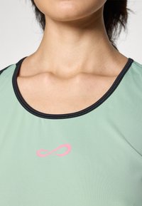 En tætsiddende atletisk shirt i mintgrøn med en sort halsudskæring. Har et pink uendelighedslogo på fronten. Glat, åndbart stof.